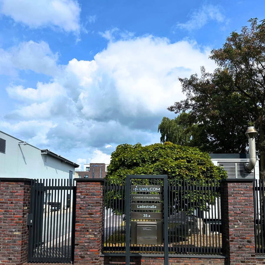 Seitenansicht des mehrstöckigen Firmengebäudes von LWLcom aus dunkelroten Ziegeln mit mehreren Fenstern und dem Firmenlogo unten rechts auf hellblauem Hintergrund.