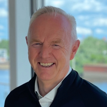  Blair Wood, Business Developer bei LWLcom. Er lacht und trägt ein weißes Hemd und eine dunkle Jacke, vor einem Fenster mit unscharfem Außenblick.