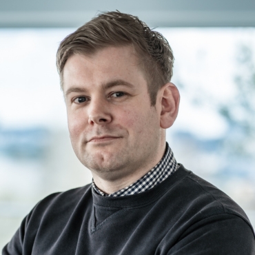 Portraitfoto von Christoph Müller, Leiter Finanzen und Einkauf bei LWLcom, trägt ein karriertes Hemd unter einem dunkelgrauen Pullover, steht vor einem Fenster mit unscharfem Hintergrund. 
